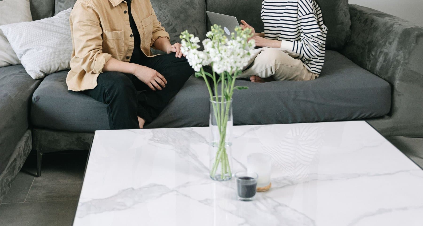 Two people talking on a couch in a living room, a laptop open between them